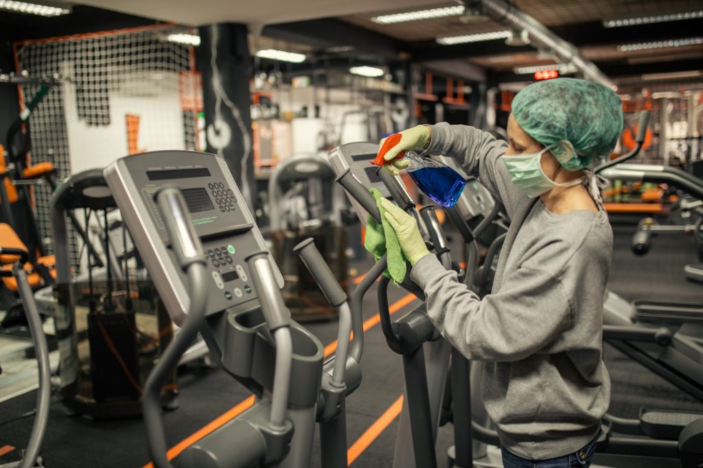 Medewerker met mondmasker desinfecteert fitnessapparatuur in sportcentrum voor een hygiënische sportomgeving