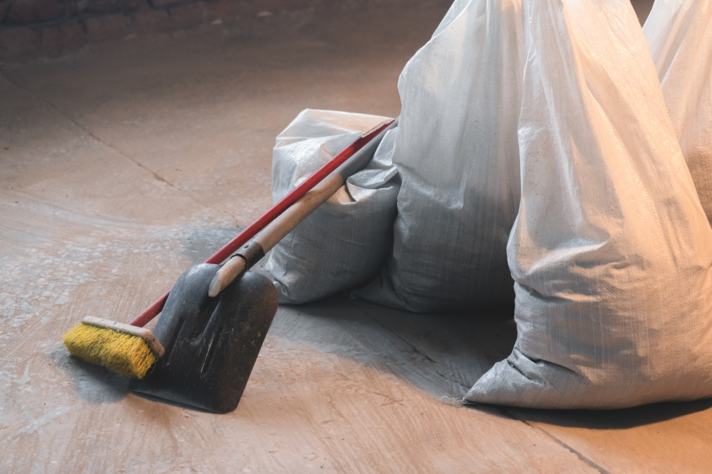 Zakken met bouwafval en gereedschap voor schoonmaak na verbouwing, inclusief schop en bezem op stoffige vloer