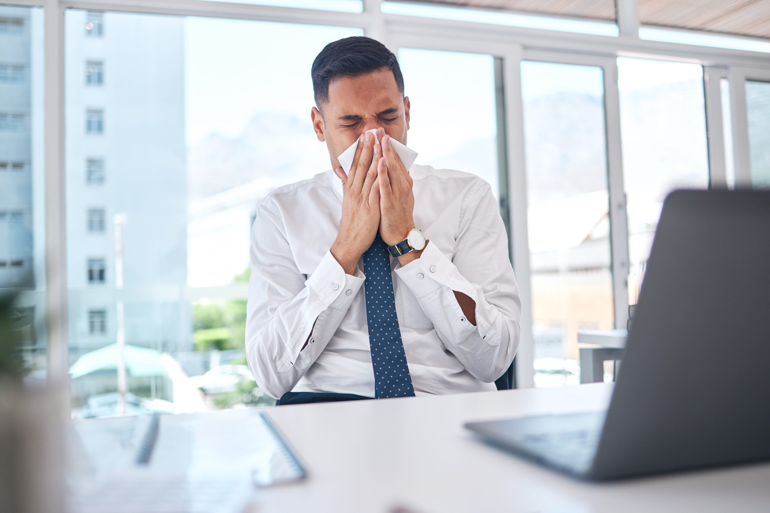 Man niest op kantoor aan zijn bureau — allergische reactie op stof of pollen op de werkvloer